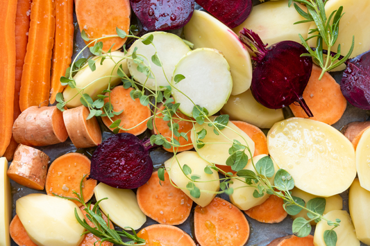 Image of fresh carrots, sweet potatoes, beets, and potatoes on a cutting board with basil and rosemary on top.