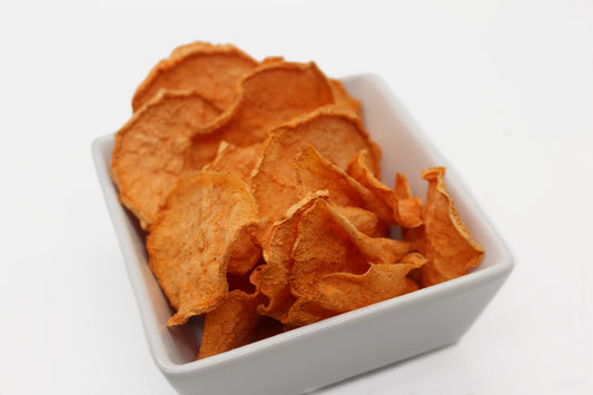 A square, white bowl filled with dehydrated sweet potato chips lightly tossed in cinnamon. The chips are a beautiful bright orange color with flecks of cinnamon.
