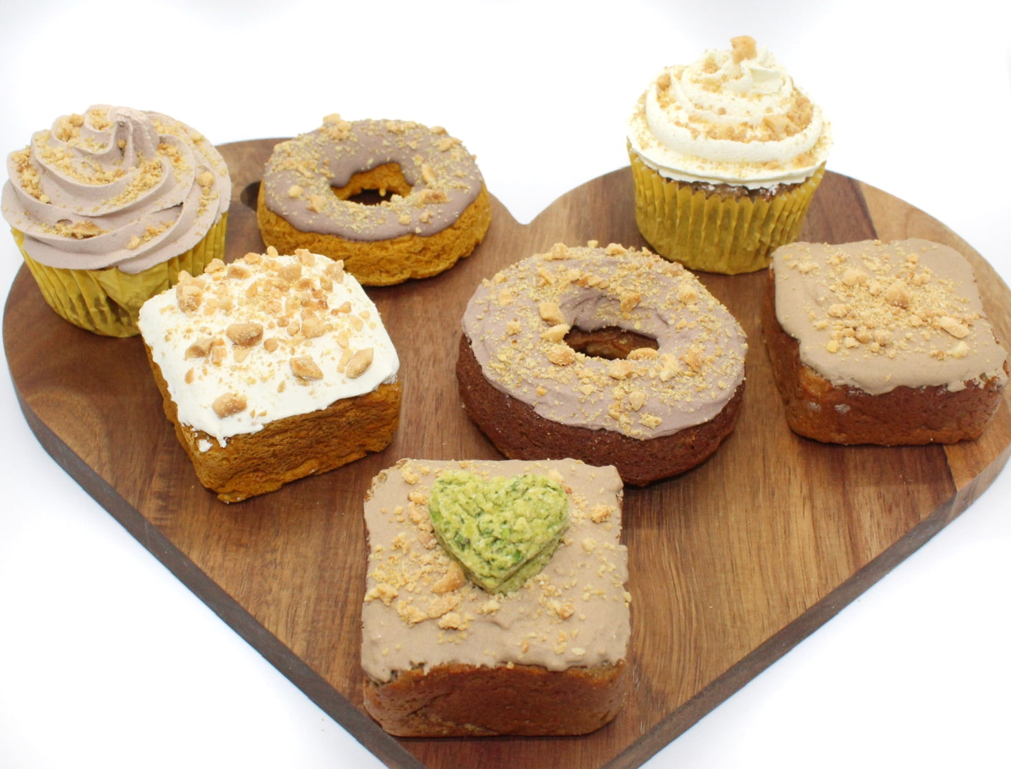 Seven various dog treats are on a wood heart-shaped cutting board. Two of the dog treats are Carob frosted donuts. The two dog cupcakes have plain and carob frosting. Three treats are dog cupcakes, two have carob frosting and the other is plain. All dog treats are sprinkled with flaxseed and crushed peanuts.