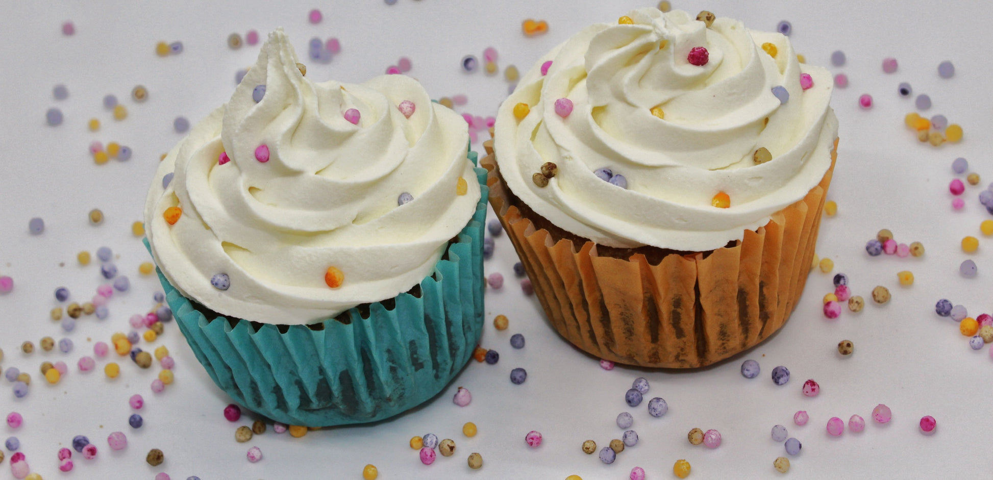 Two colorful cupcakes each wrapped in blue and orange paper liner with white swirled frosting and rainbow pearl sprinkles on top. There are rainbow pearl sprinkles scattered throughout the background.