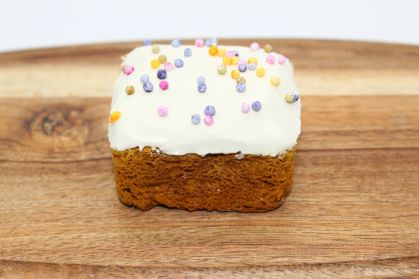 A sweet potato and carrot mini dog cake with white frosting and rainbow pearl sprinkles on top which is displayed on a wood cutting board.