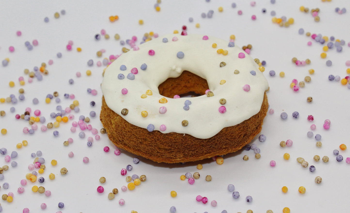 A apple and banana flavored dog donut with white frosting and dog safe rainbow pearl sprinkles on top. There are rainbow pearl sprinkles scattered in the background.