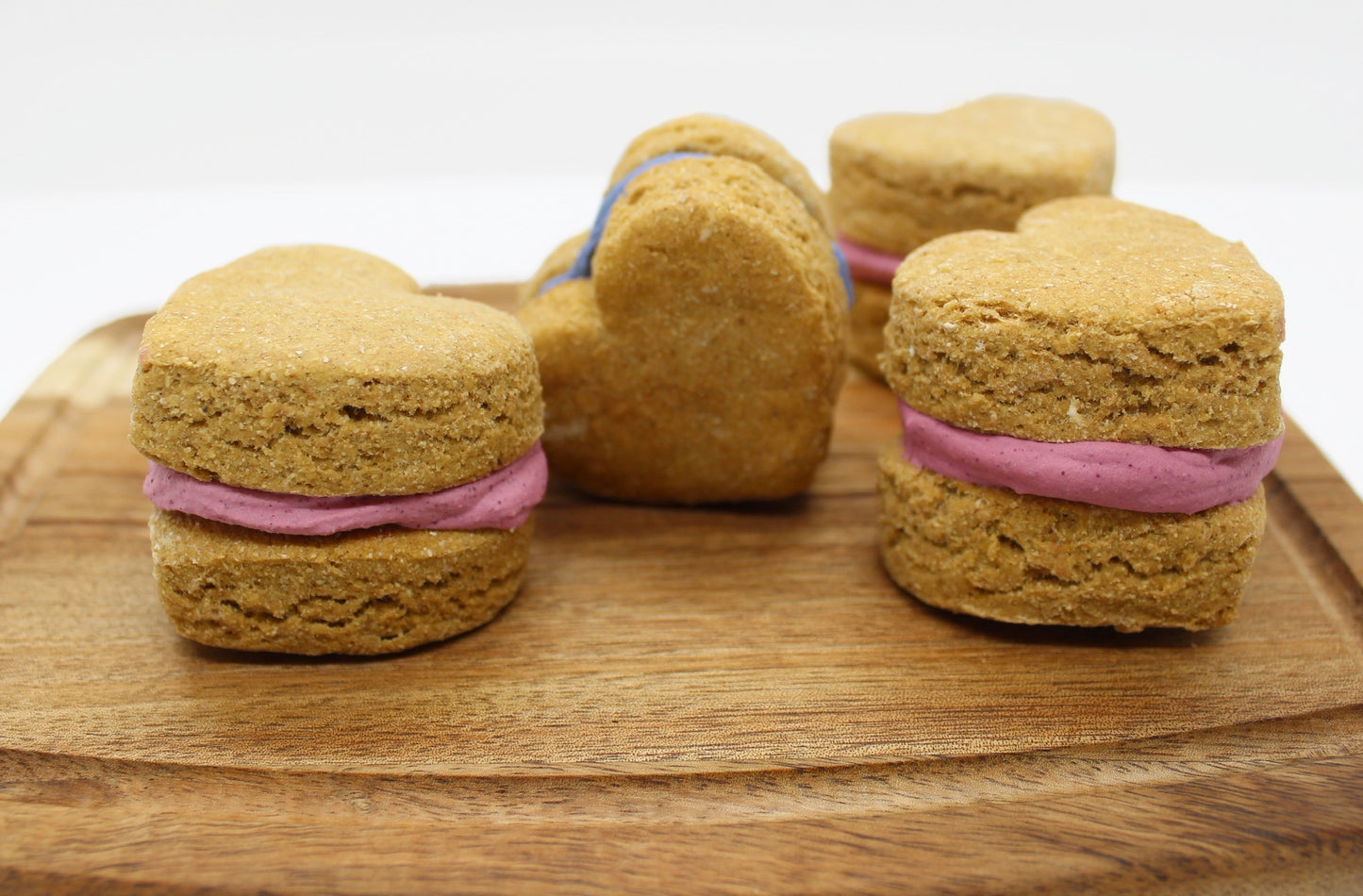 Four apple and banana flavored whoopie hearts on a cutting board. Each whoopie heart has two cookies with icing sandwiched in the middle. Three whoopie hearts have pink icing in the middle and one has blue icing.
