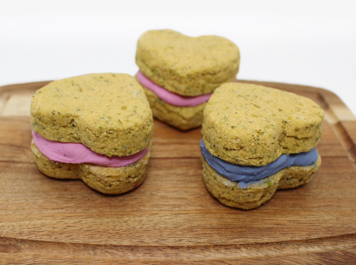 Three Broccoli and Peas whoopie heart dog treats on a cutting board. Two have pink icing and the other has blue in the middle.
