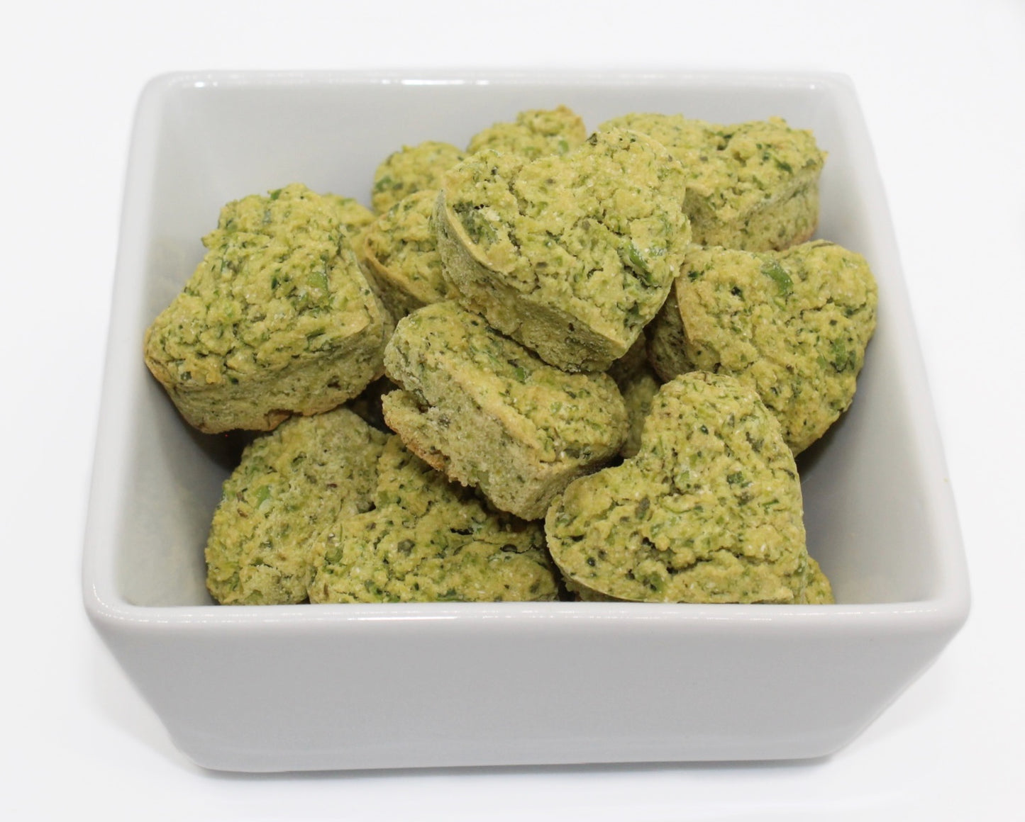 A bunch of green heart-shaped dog training treats in a white bowl. The treats are Broccoli and Peas flavor, parsley and cilantro can be seen in the treats.