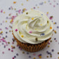 A artfully displayed cupcake viewed from an aerial shot with white, piped frosting and rainbow pearl sprinkles on top. There are rainbow pearl sprinkles scattered throughout the background.