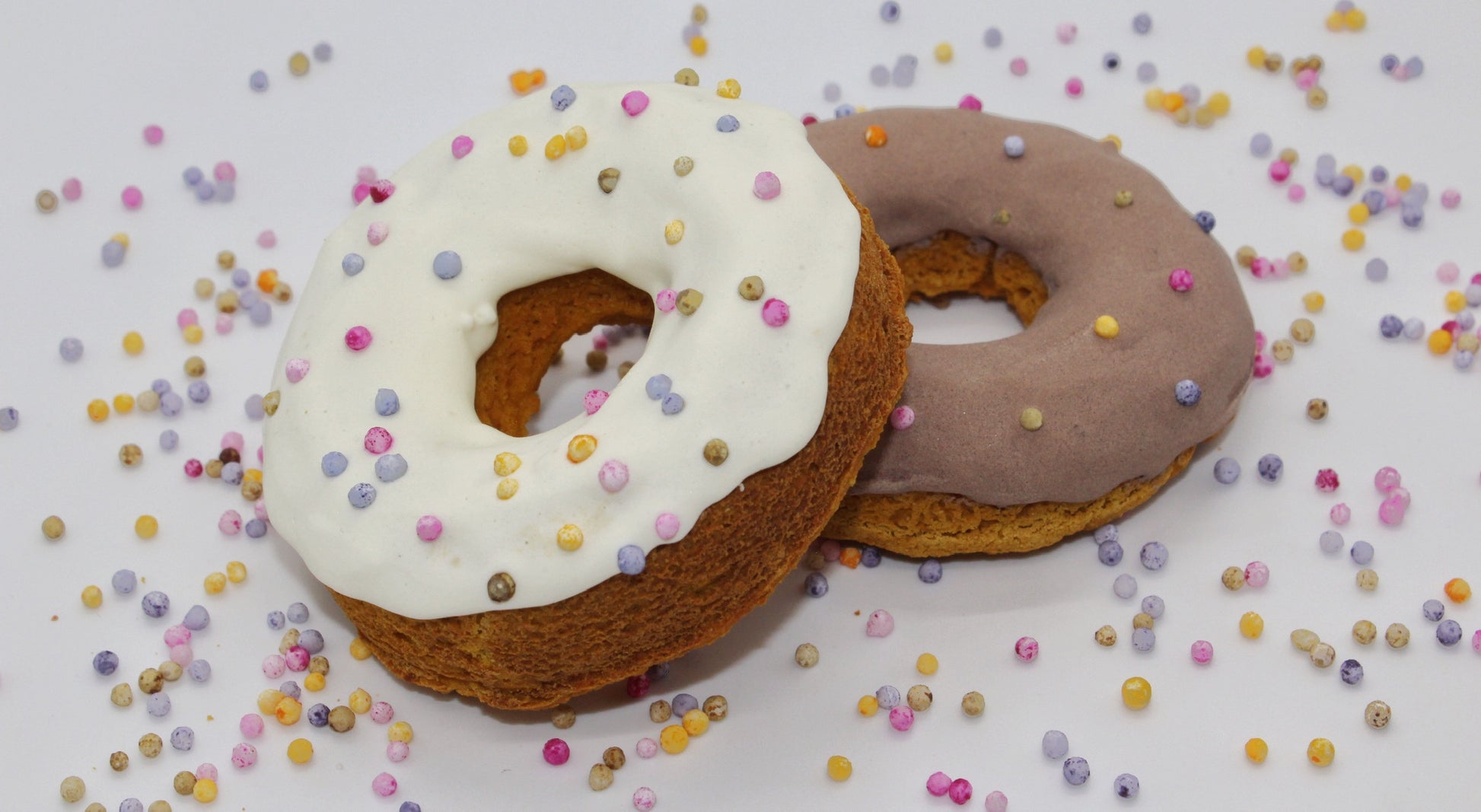 Two frosted dog donuts with dog safe rainbow pearl sprinkles on top. One donut is apple and banana flavored with white frosting. The second donut is pumpkin flavored with brown carob frosting. There are rainbow pearl sprinkles scattered in the background.
