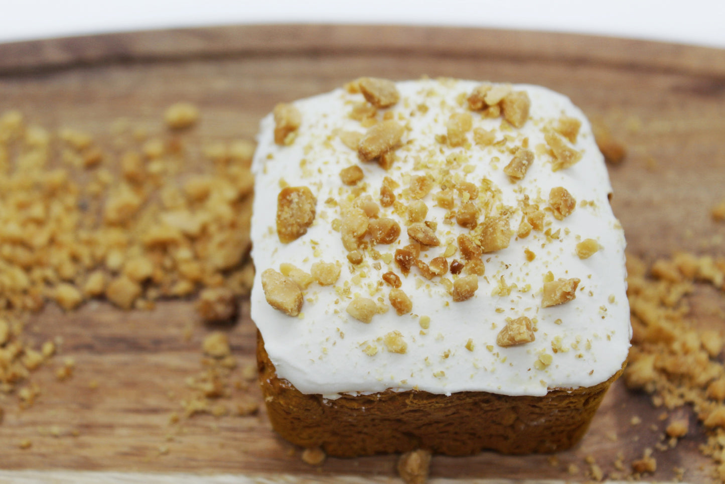 A beautiful sweet potato and carrot flavored mini dog cake with white frosting, peanuts and flaxseed sprinkled on top. The mini cake is displayed on a small cutting board with peanuts and flaxseed sprinkled all around.