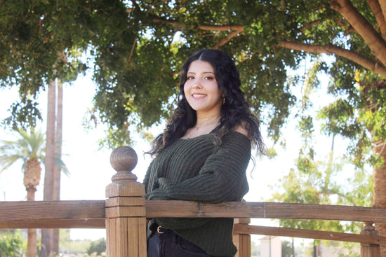 A nice photo of owner Alyssa standing on a bridge under a tree. Alyssa is wearing a green sweater and black denim pants. Alyssa's hair is curly and she is wearing gold hoop earrings.