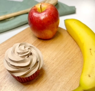 A delicious dog cupcake is placed on a wood cutting board next to a fresh apple and banana.