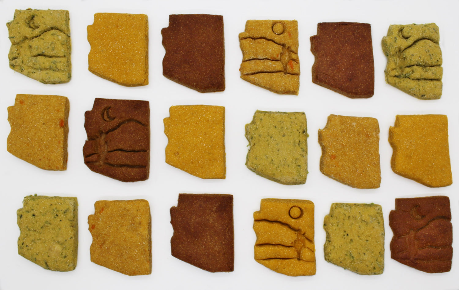 18 dog biscuits are placed in three rows of six on a white surface. The dog biscuits are all cut in the shape of the state of Arizona. Six of the biscuits have a desert motif stamped into them. The biscuits are placed randomly with the flavors of biscuits ranging from Broccoli and Peas, Sweet potato and Carrots, Pumpkin, and Peanut butter and Banana.