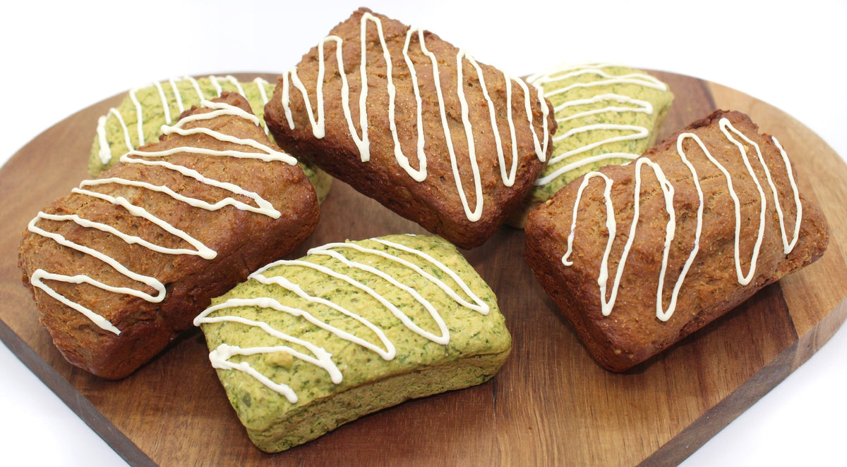 Six loaves with a drizzle of frosting on a heart-shaped cutting board. Three of the loaves are Broccoli and Peas, the other three are Peanut butter and Banana.