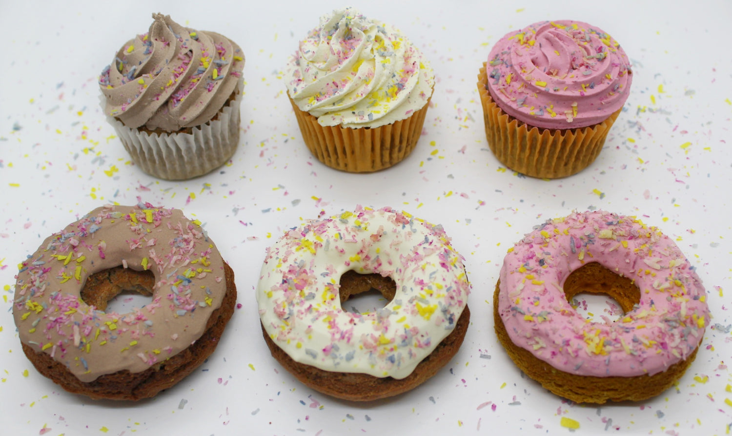 Six delicious bakery items covered in rainbow coconut shreds. The front row has three differently frosted donuts carob brown, plain white, and beet pink. The back row has three frosted dog cupcakes, there is carob brown, plain white, and beet pink.