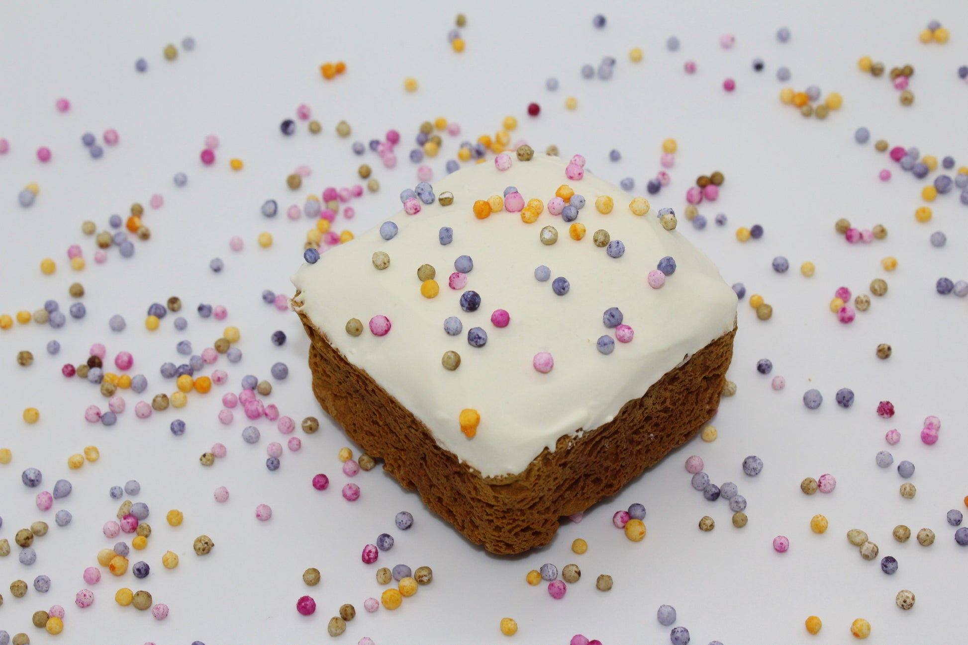 A sweet potato and carrot mini dog cake with white frosting and rainbow pearl sprinkles on top. There are rainbow pearl sprinkles all over the background.