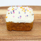 A sweet potato and carrot mini dog cake with white frosting and rainbow pearl sprinkles on top which is displayed on a wood cutting board.