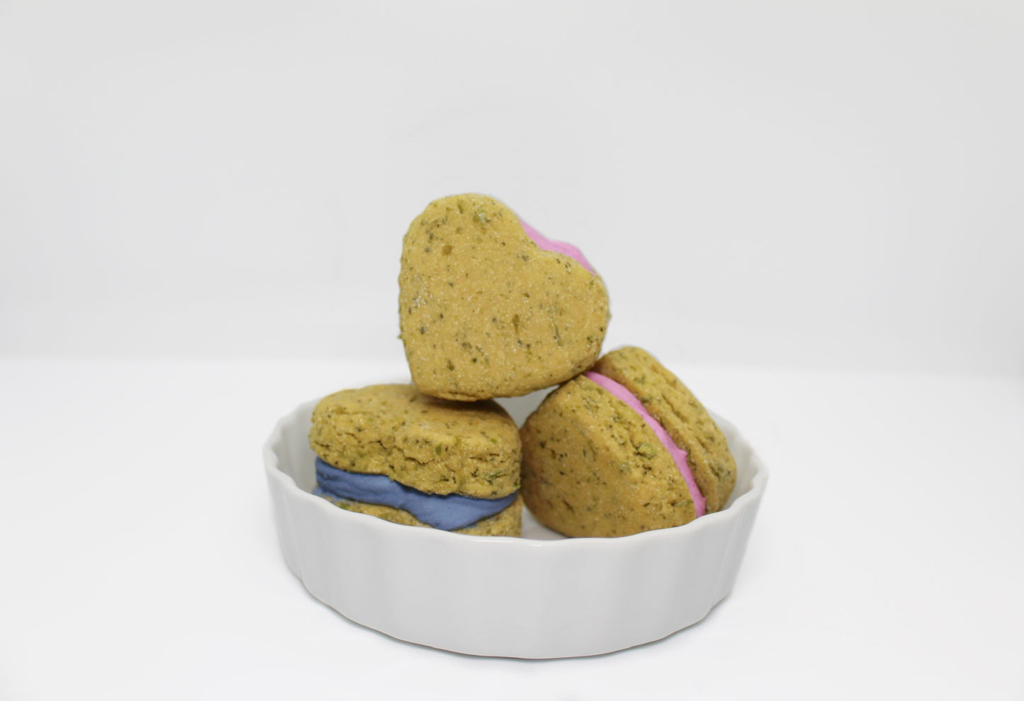Three green whoopie heart dog treats in a white dish. The whoopie consists of two Broccoli and Peas cookies with frosting sandwiched in the middle. The frosting colors displayed are  pink and blue.