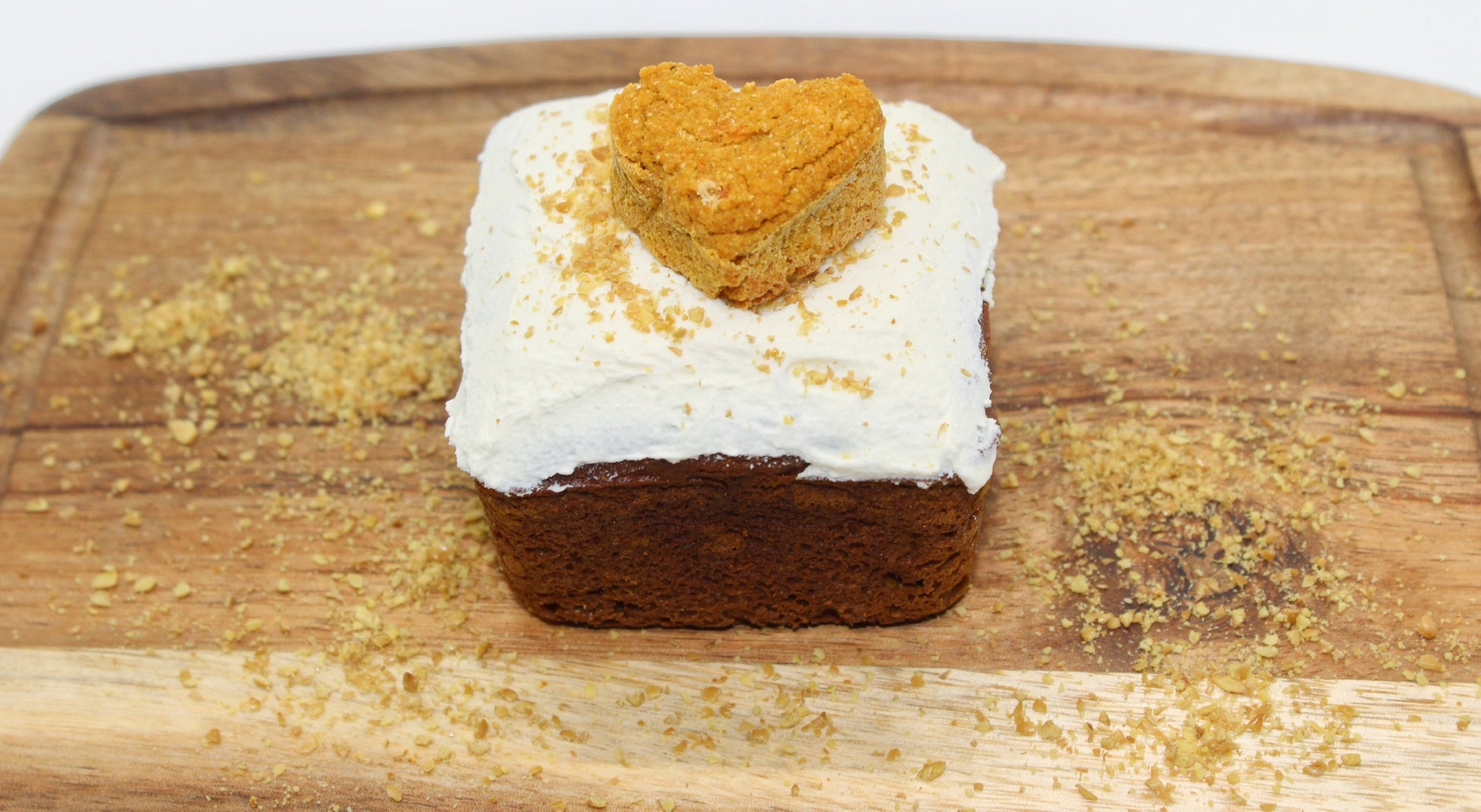 A beautiful sweet potato and carrot flavored mini dog cake with white frosting and flaxseed sprinkled on top. This yummy mini cake also has a heart shaped sweet potato and carrot training treat on top. The mini cake is displayed on a small cutting board with flaxseed sprinkled all around.