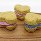 Three Broccoli and Peas whoopie heart dog treats on a cutting board. Two have pink icing and the other has blue in the middle.