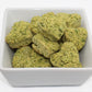A bunch of green heart-shaped dog training treats in a white bowl. The treats are Broccoli and Peas flavor, parsley and cilantro can be seen in the treats. 