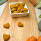 A white, square bowl filled with bite-sized, heart-shaped training treats in sweet potato and carrot flavor. The bowl is on a cutting board with two fresh sweet potatoes and two whole carrots in the background.