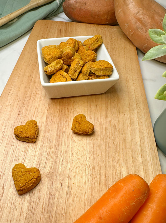 A white, square bowl filled with bite-sized, heart-shaped training treats in sweet potato and carrot flavor. The bowl is on a cutting board with two fresh sweet potatoes and two whole carrots in the background.