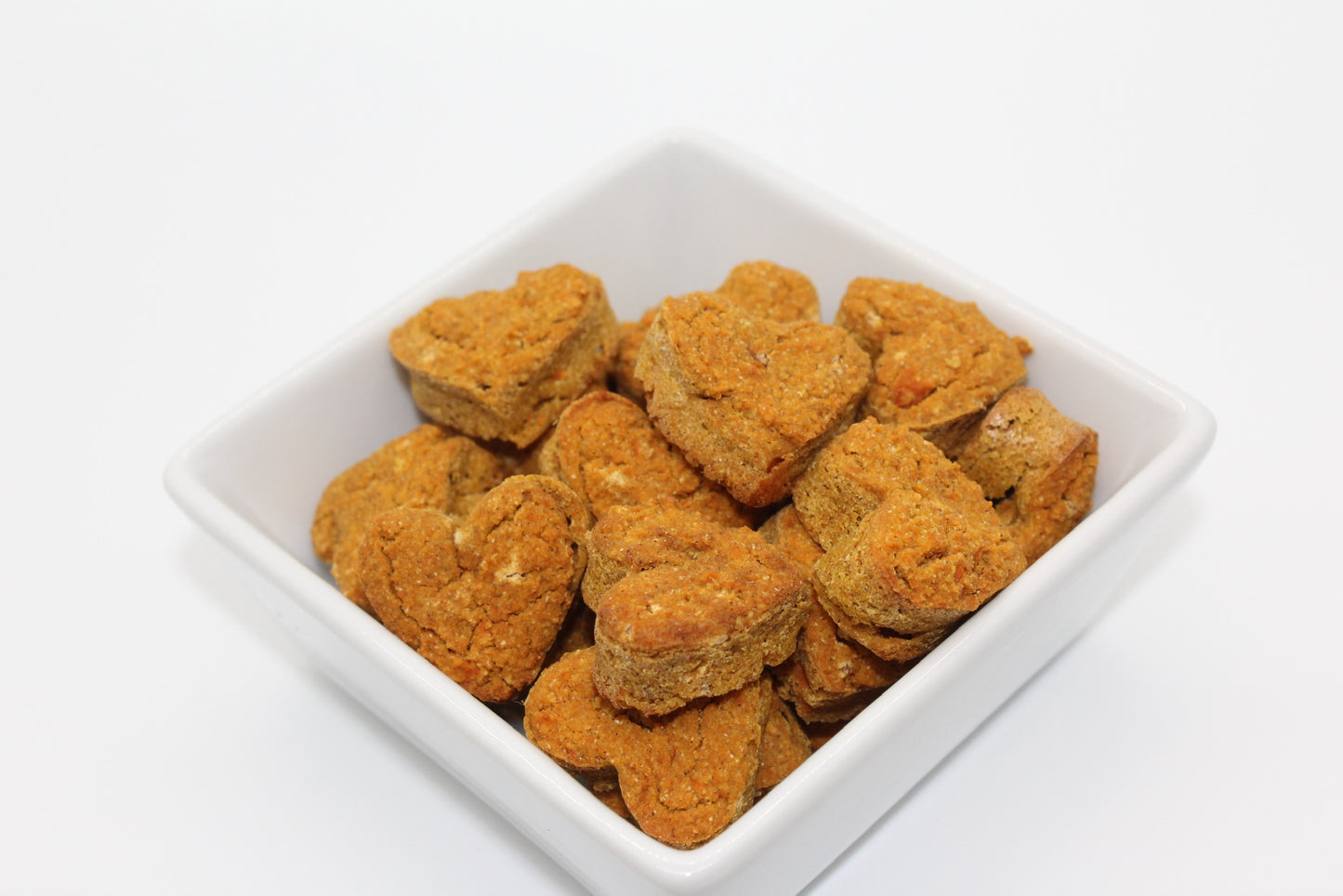 A white, square bowl filled with bite-sized, heart-shaped training treats for dogs in sweet potato and carrot flavor. Pieces of carrots can be seen throughout the treats.