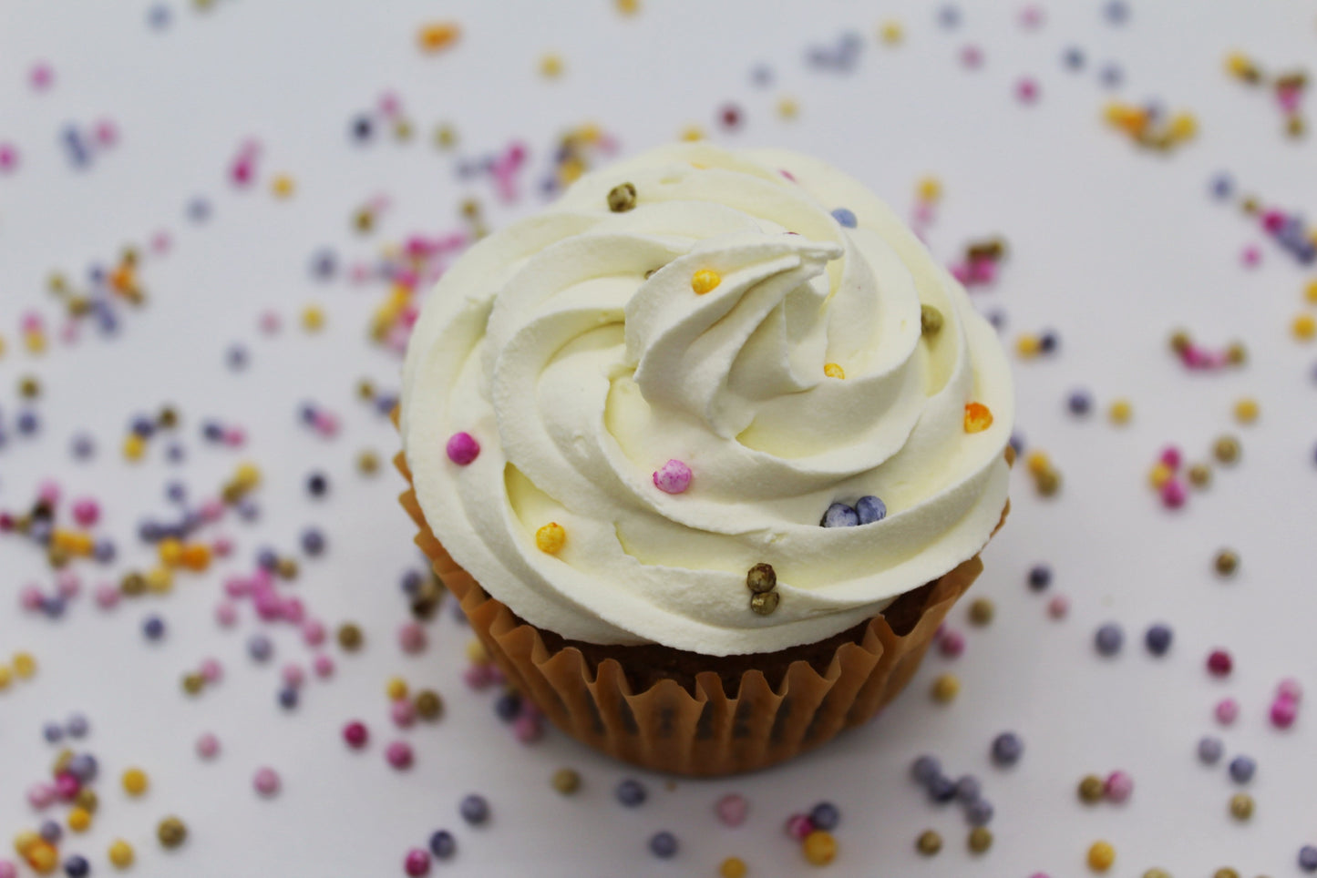 A artfully displayed cupcake viewed from an aerial shot with white, piped frosting and rainbow pearl sprinkles on top. There are rainbow pearl sprinkles scattered throughout the background.