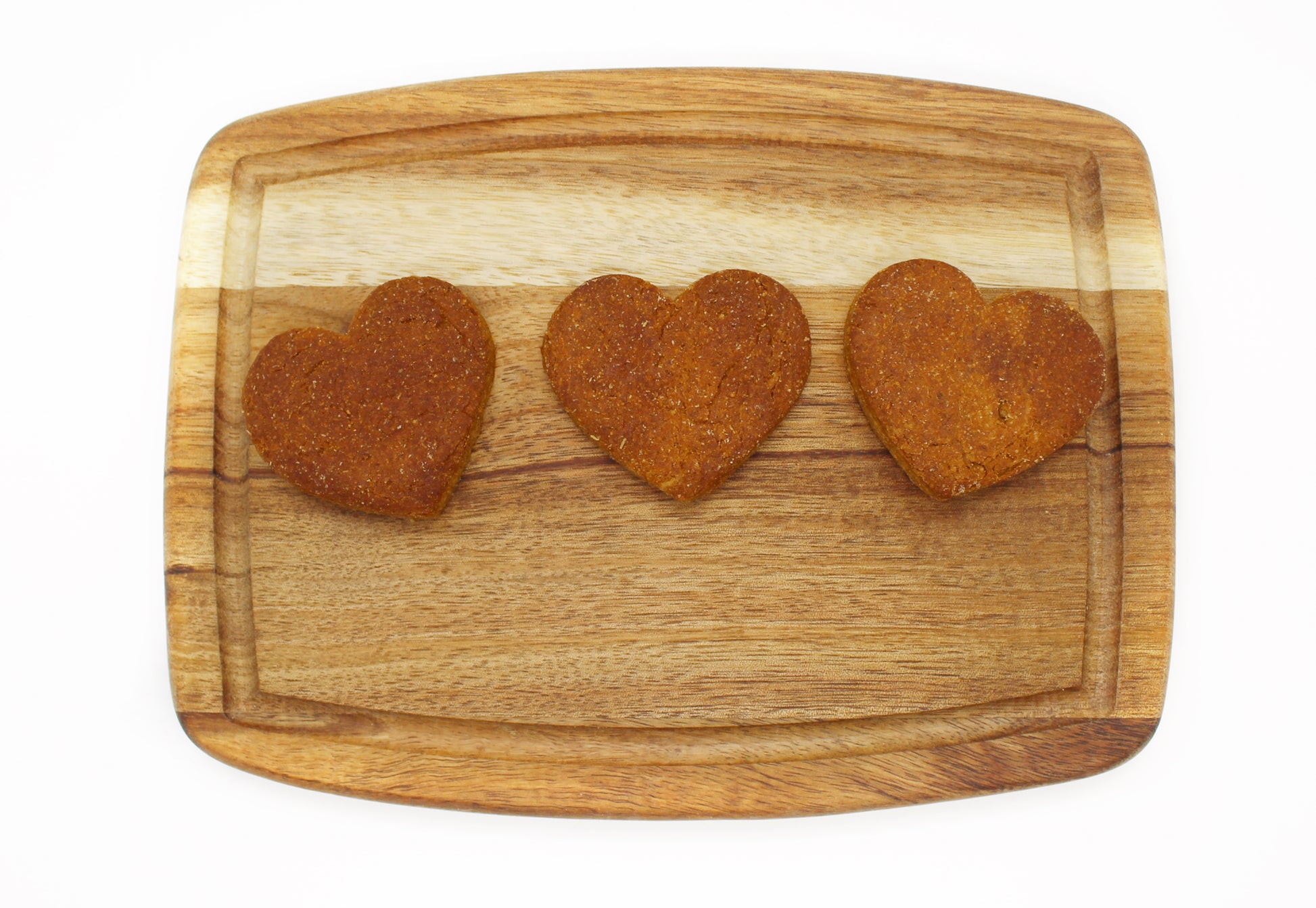 Three heart-shaped peanut butter and banana flavored dog biscuits laid out in a single line on a cutting board.