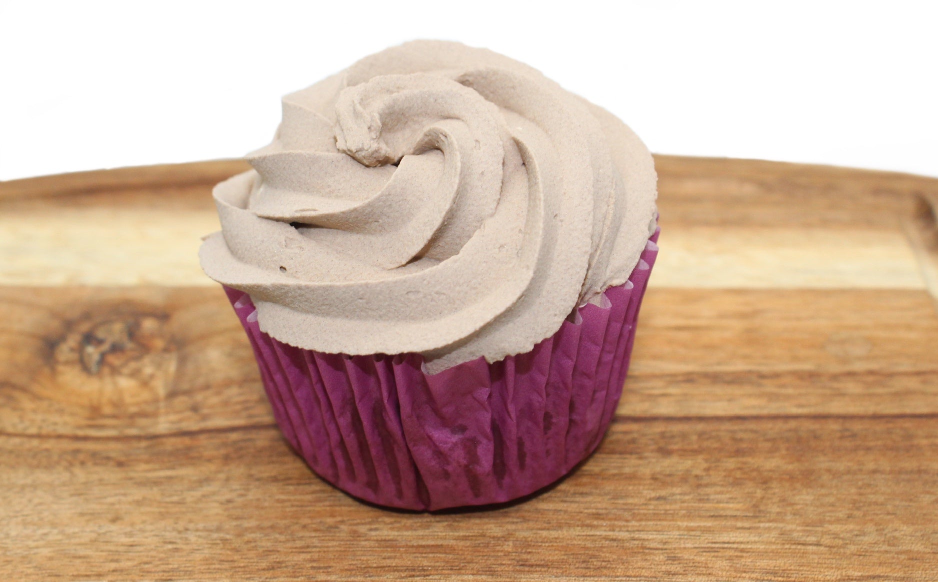 A cupcake wrapped in a purple paper liner with brown, carob frosting. The cupcake is displayed on a wood cutting board.