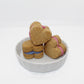 Three apple and banana flavored whoopie hearts displayed in a white dish. Two whoopie hearts have pink icing in the middle and one has blue icing.