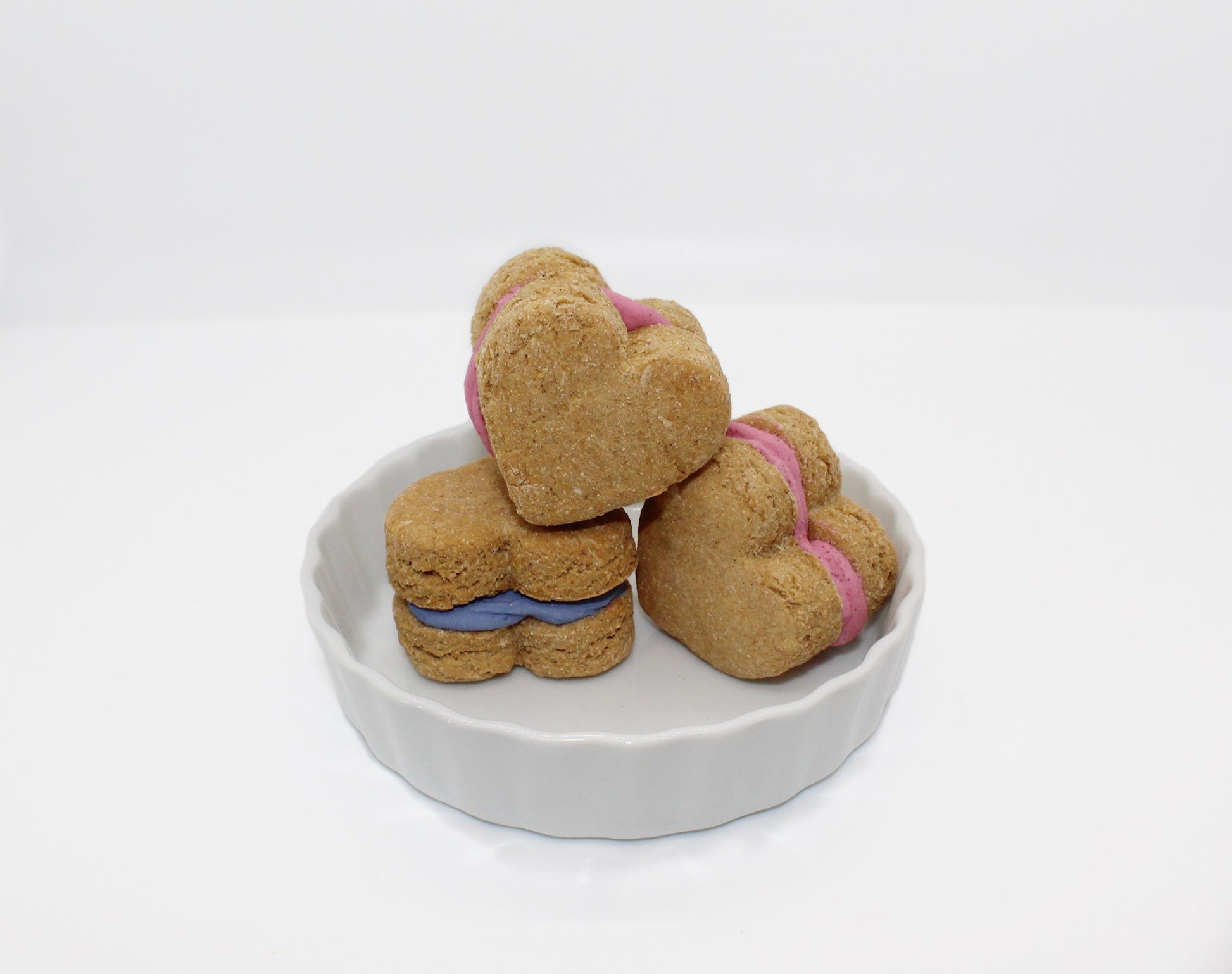 Three apple and banana flavored whoopie hearts displayed in a white dish. Two whoopie hearts have pink icing in the middle and one has blue icing.
