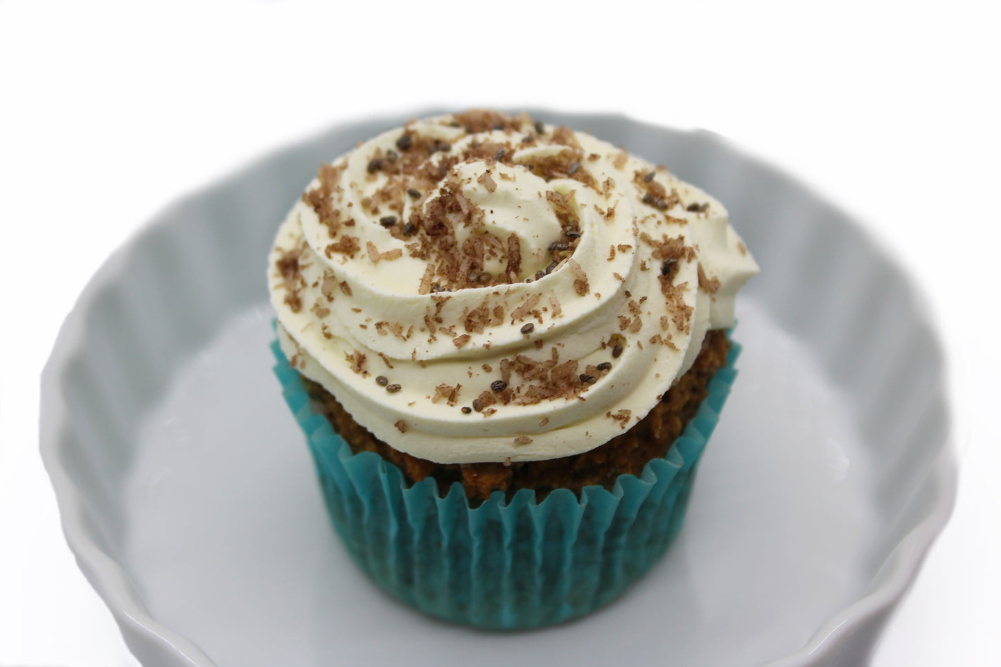 A cupcake wrapped in a blue paper liner with white frosting piped on top and brown, carob powdered coconut flakes displayed in a round, white dish.