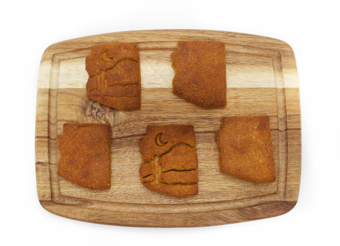 Five peanut butter and banana flavor dog biscuits cut in the shape of the state of Arizona on a cutting board. Two of the biscuits have a desert themed stamp of a moon, mountains, and a cactus.