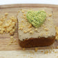 A mini dog cake with brown, carob frosting, peanuts and  flaxseed sprinkled on top. This yummy mini cake also has a heart shaped broccoli and peas training treat on top. The mini cake is displayed on a small cutting board with crushed peanuts and flaxseed sprinkled all around.