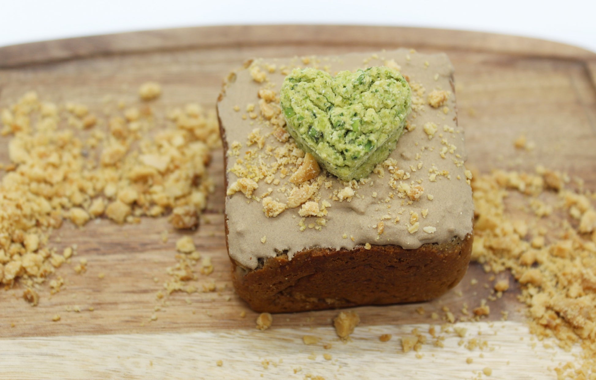 A mini dog cake with brown, carob frosting, peanuts and  flaxseed sprinkled on top. This yummy mini cake also has a heart shaped broccoli and peas training treat on top. The mini cake is displayed on a small cutting board with crushed peanuts and flaxseed sprinkled all around.