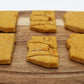 Six sweet potato and carrot flavored dog biscuits in the shape of the state of Arizona displayed on a cutting board. Two biscuits have a stamp of a desert scene which show a sun, cactus and mountains. 