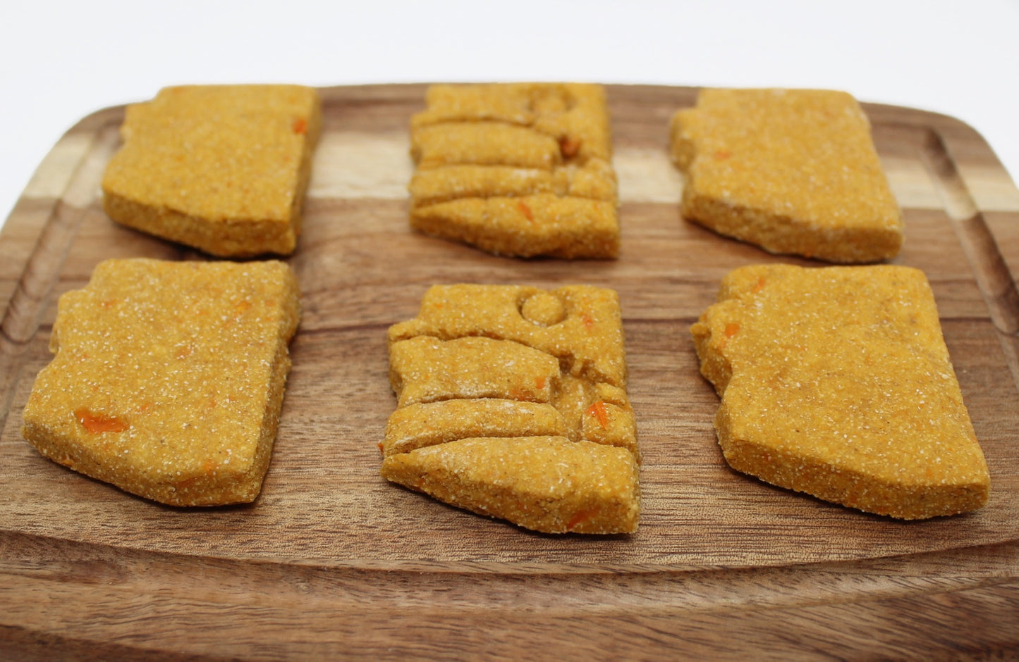 Six sweet potato and carrot flavored dog biscuits in the shape of the state of Arizona displayed on a cutting board. Two biscuits have a stamp of a desert scene which show a sun, cactus and mountains. 